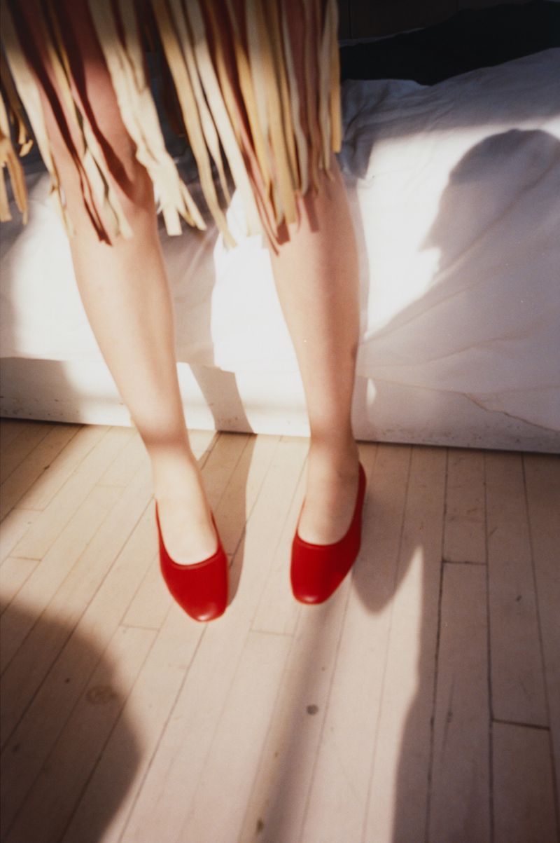 A woman is sitting with red flats on a wooden floor in front of a white bed, illuminated by sunlight and shadows.