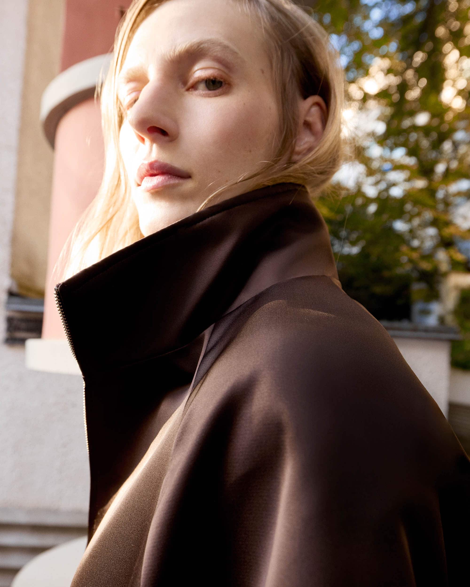 Close-up portrait of a fair-skinned woman with blonde hair wearing a brown top against a blurred architectural and foliage background.