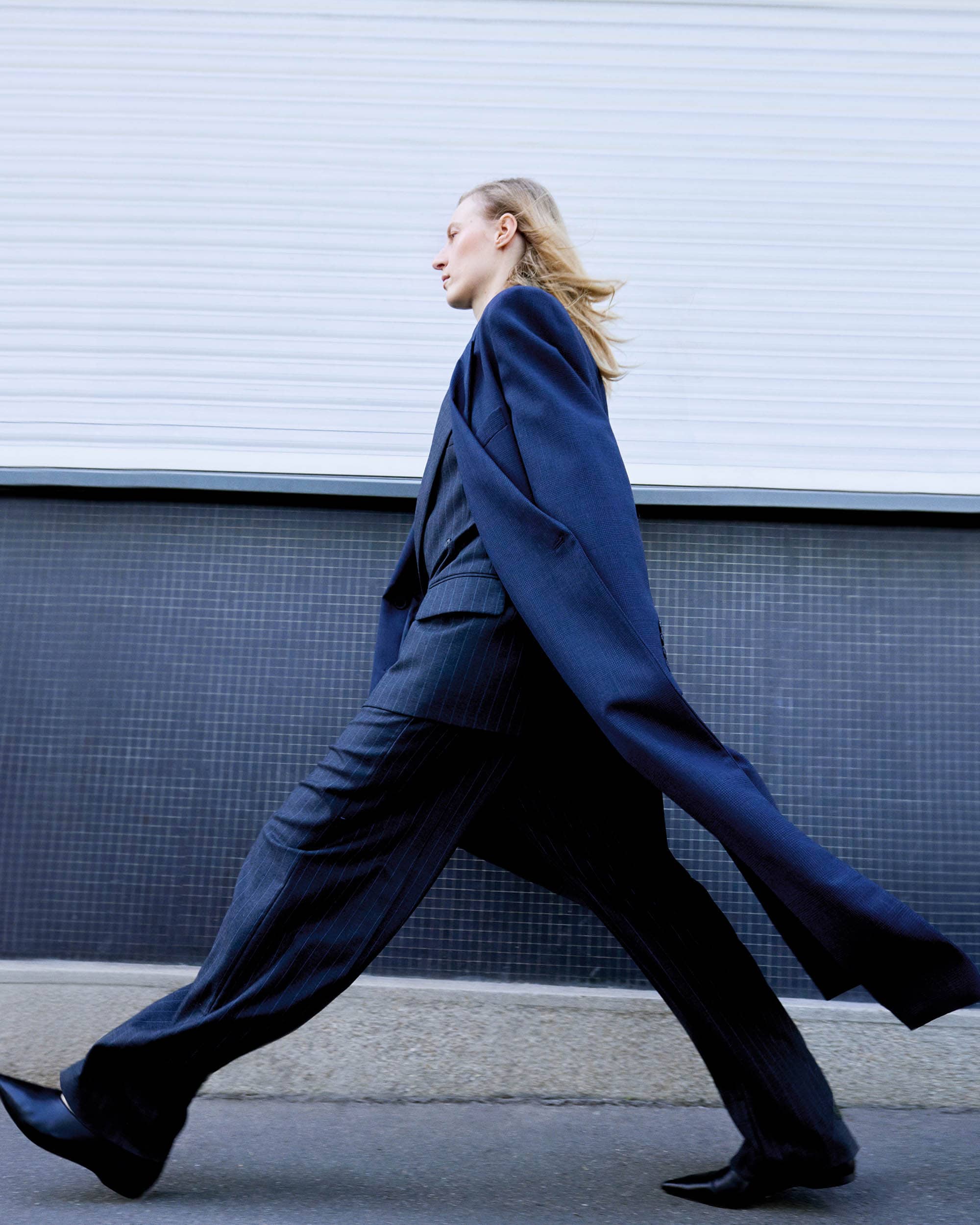 A slender woman in a dark suit strides confidently against a textured blue wall, her blonde hair catching the light.