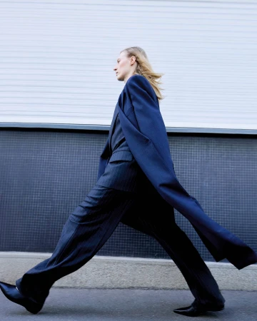 A slender woman in a dark suit strides confidently against a textured blue wall, her blonde hair catching the light.