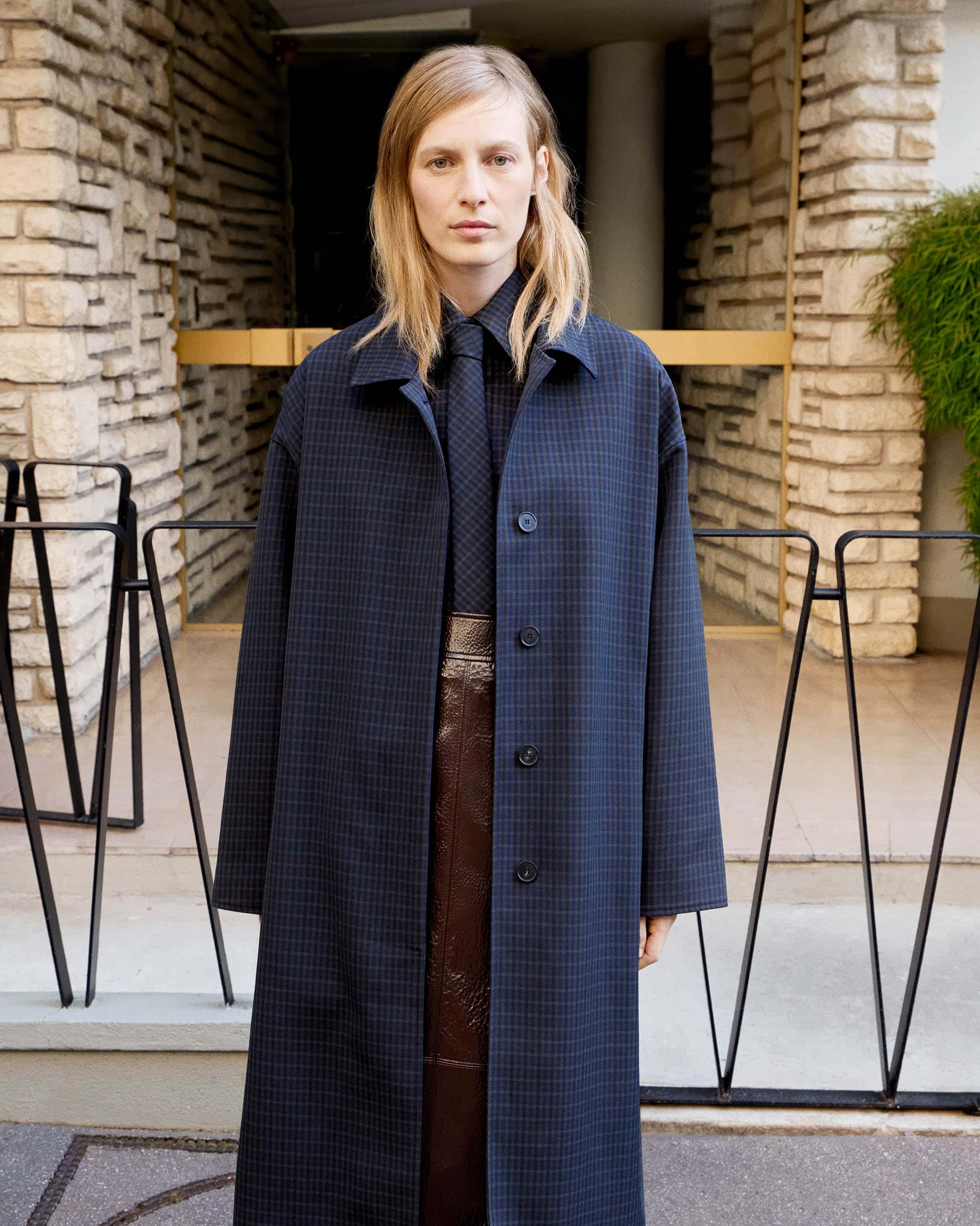 Adult woman wearing a navy checkered coat and tie stands in front of a neutral stone building with black railings. Fashion shot.