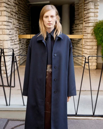 Adult woman wearing a navy checkered coat and tie stands in front of a neutral stone building with black railings. Fashion shot.