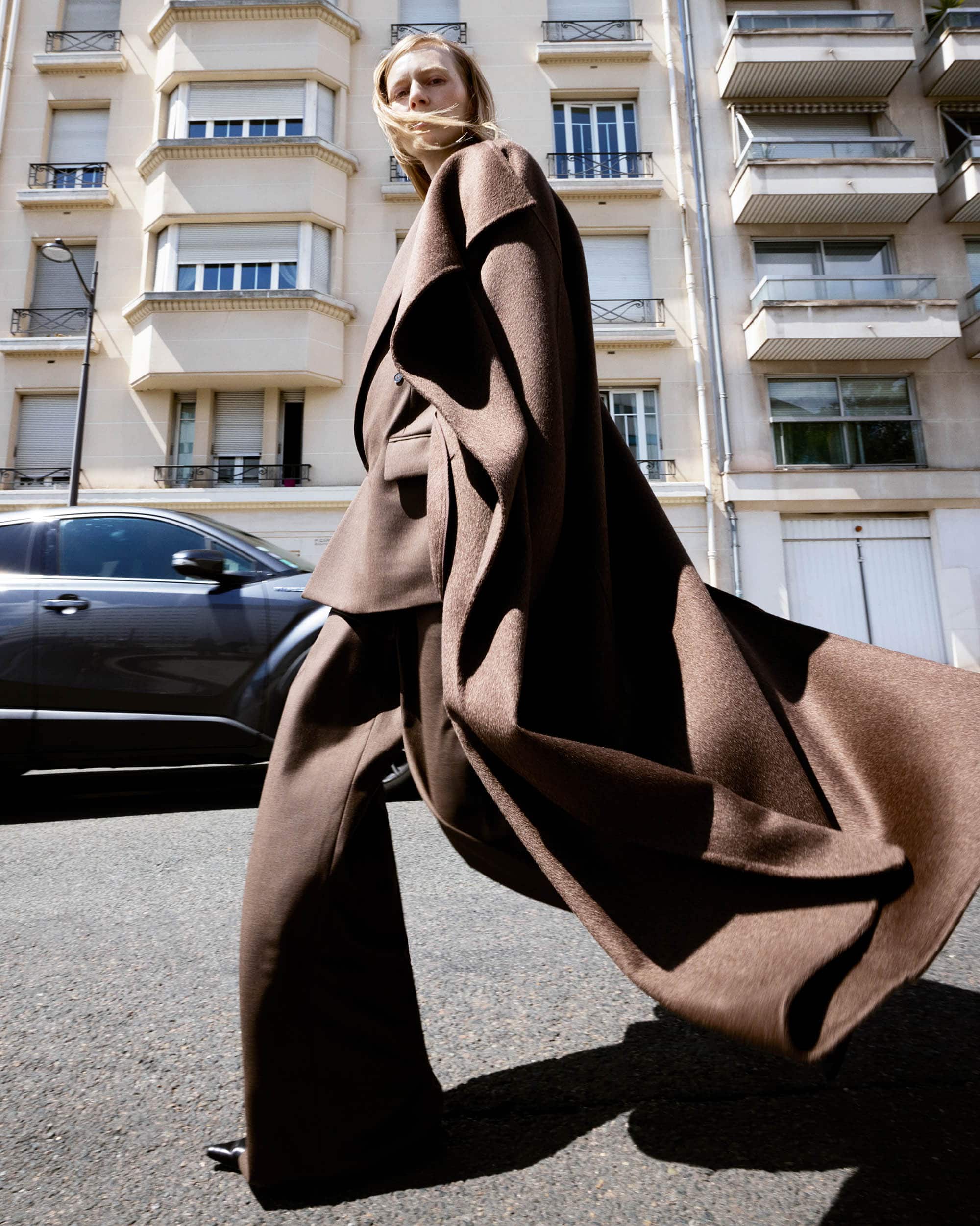 Fashionable woman in brown wide-leg suit and coat walking on a city street with parked car and building in background on a sunny day.