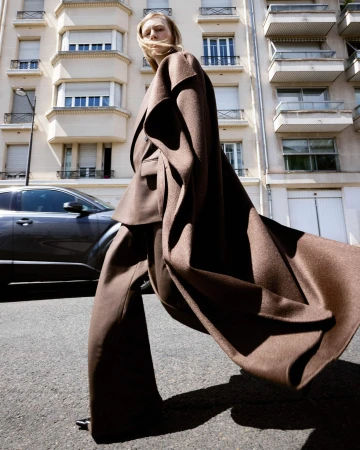 Fashionable woman in brown wide-leg suit and coat walking on a city street with parked car and building in background on a sunny day.