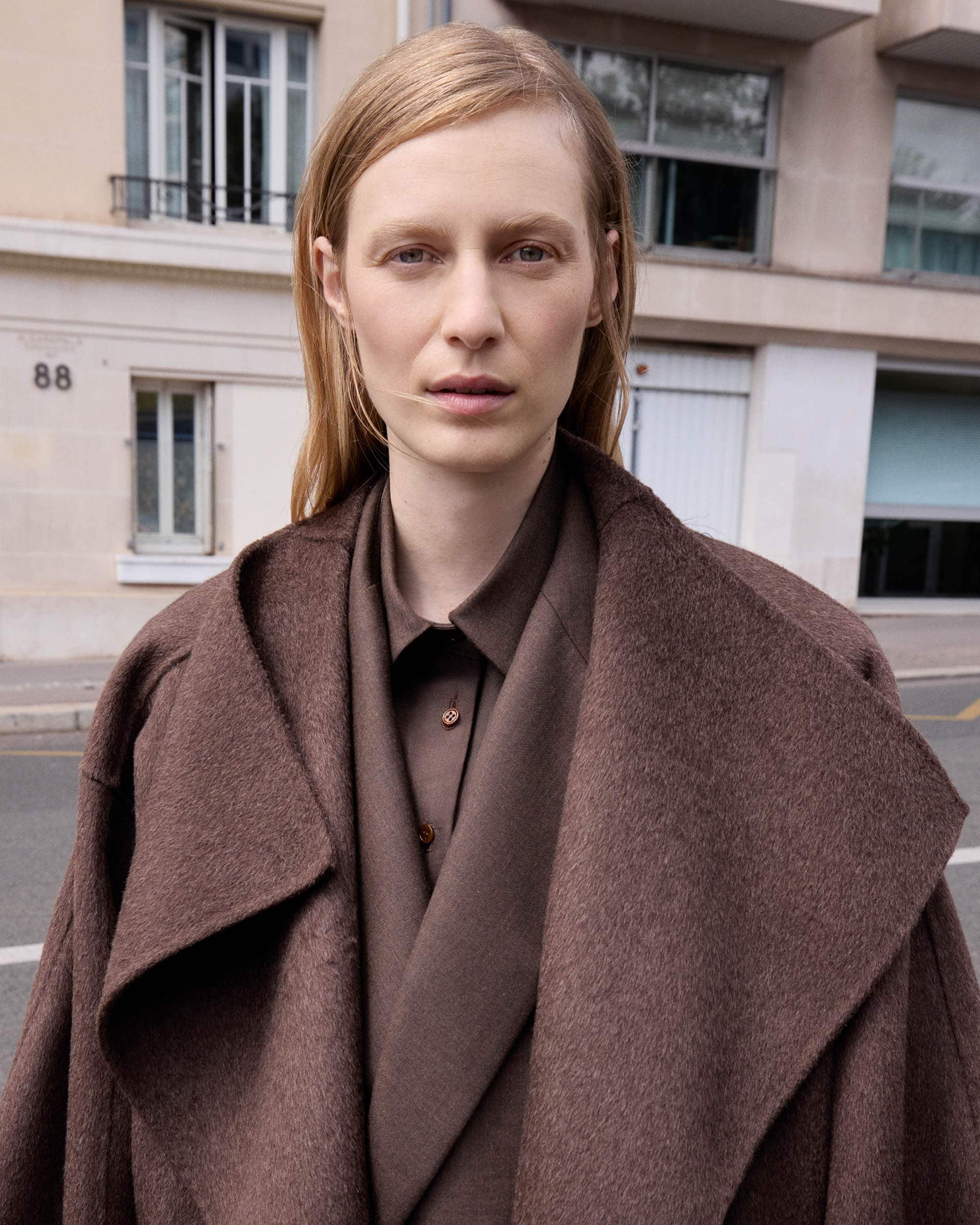 A woman with blonde hair stands outdoors wearing a brown coat and shirt, close-up shot with neutral expression and natural light.