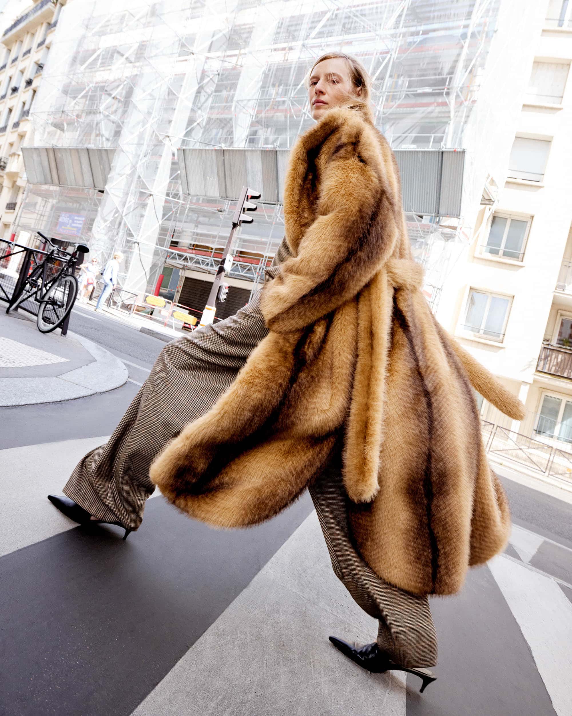 Fashionable woman in a brown fur coat strides across a crosswalk in a European city, captured in a low-angle shot with blurred background.