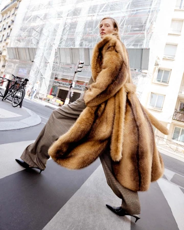 Fashionable woman in a brown fur coat strides across a crosswalk in a European city, captured in a low-angle shot with blurred background.