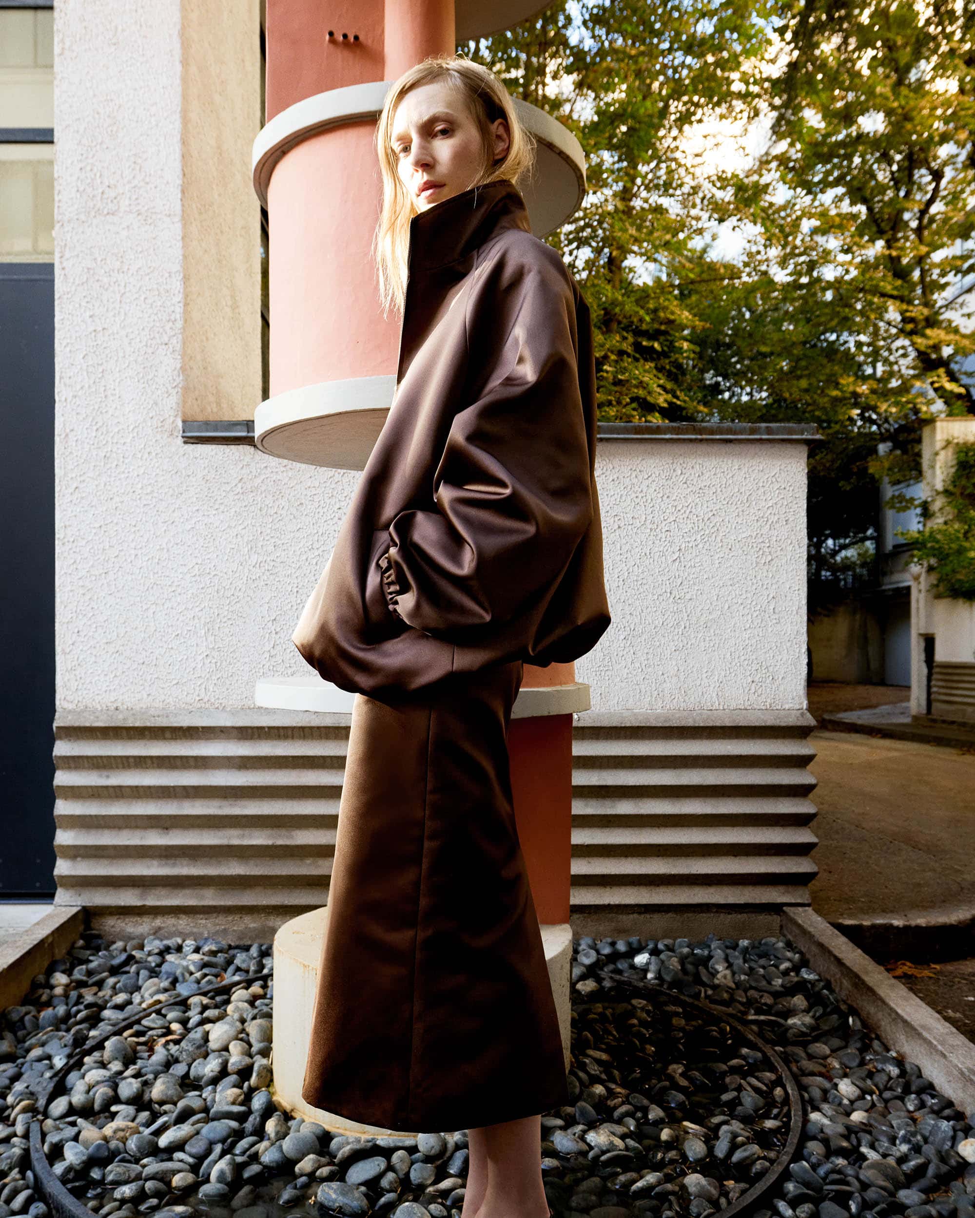 Adult female wearing a brown satin skirt suit stands outdoors against modern architecture in an eye-level shot with shallow depth of field.