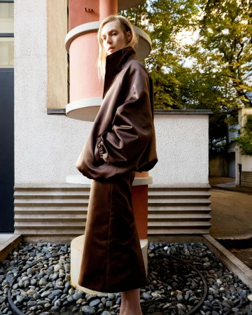 Adult female wearing a brown satin skirt suit stands outdoors against modern architecture in an eye-level shot with shallow depth of field.
