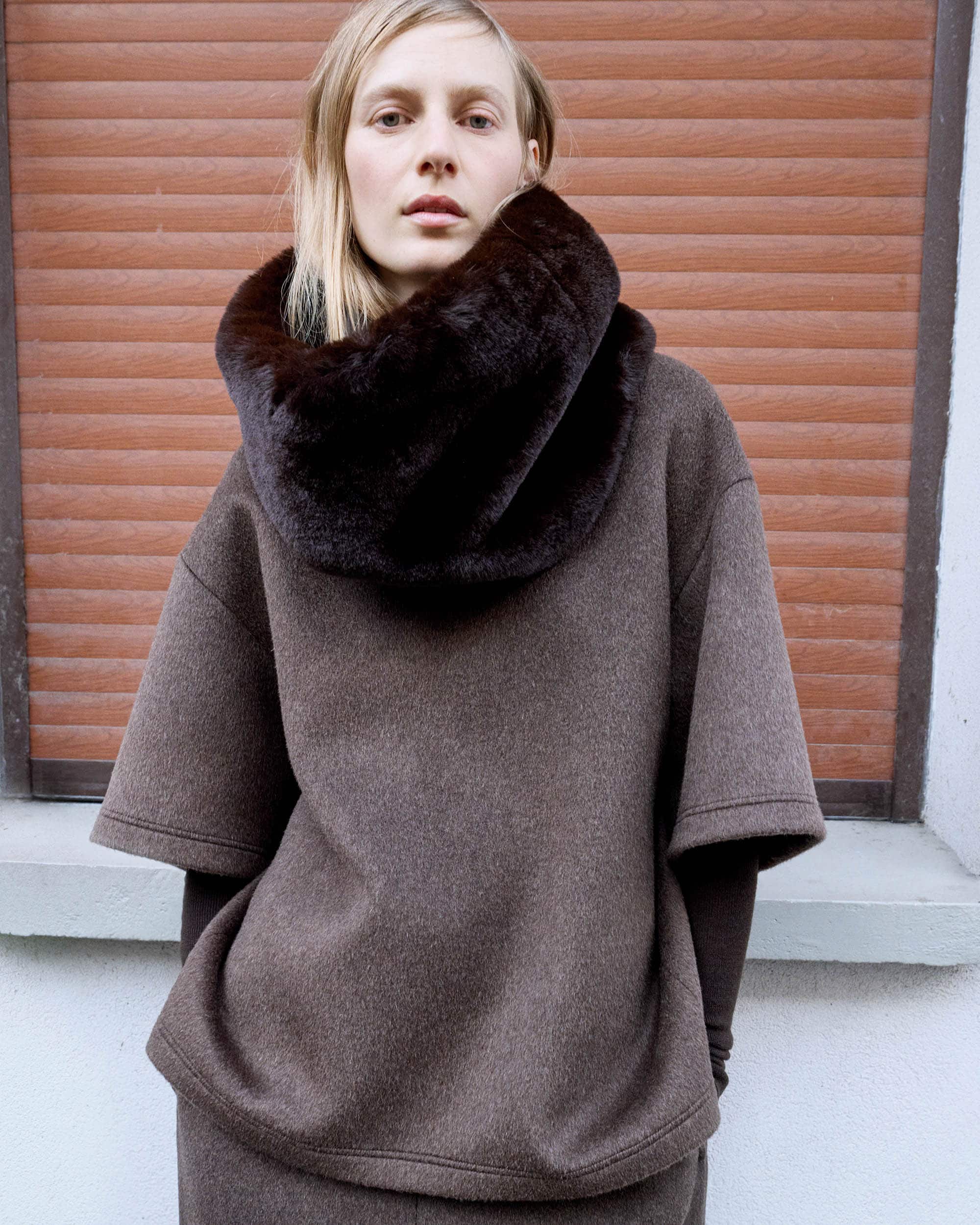 A fair-skinned woman with blonde hair stands indoors wearing a brown wool outfit with a dark brown faux fur collar. The background is a brown shutter.