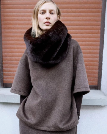 A fair-skinned woman with blonde hair stands indoors wearing a brown wool outfit with a dark brown faux fur collar. The background is a brown shutter.