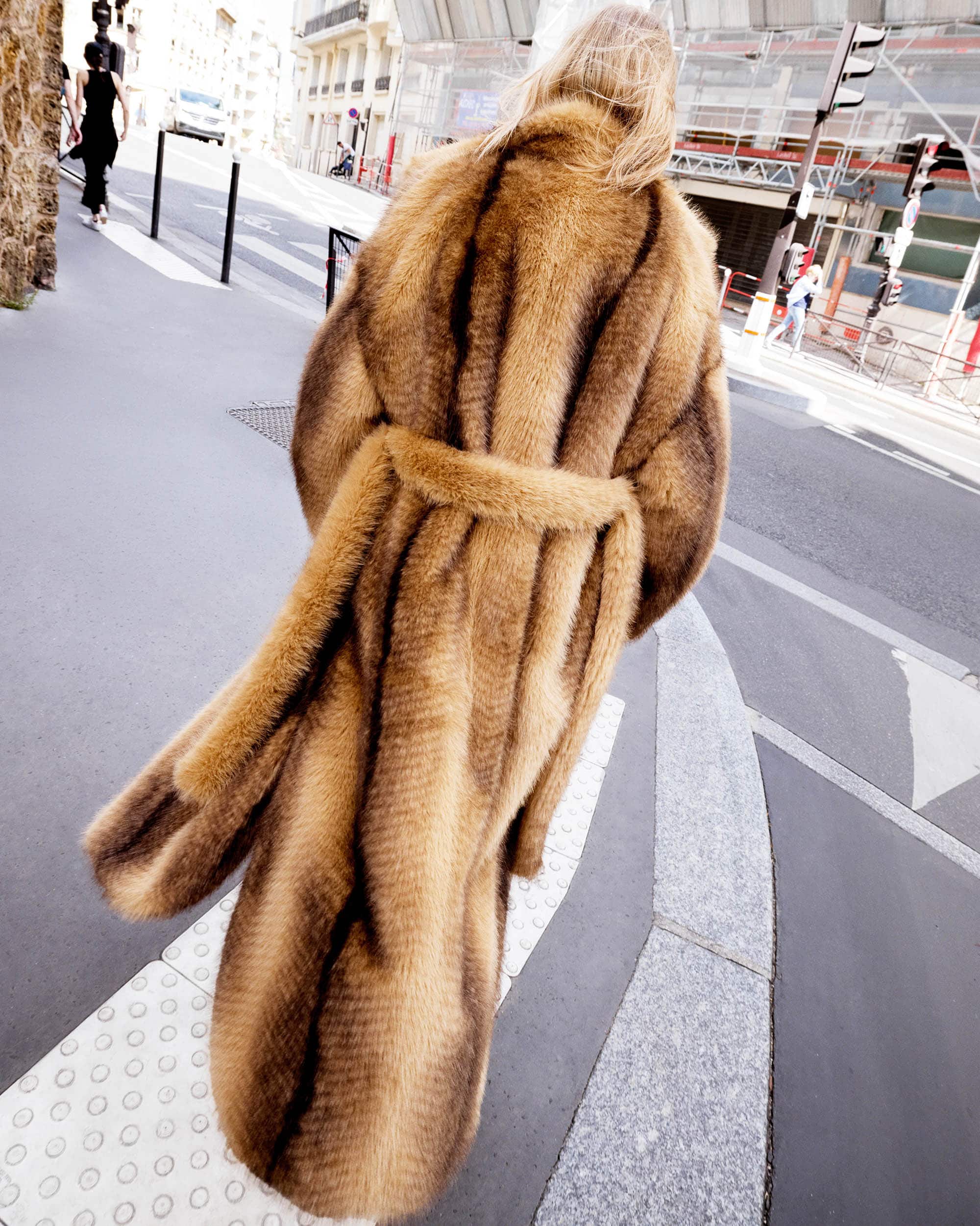A woman in a brown and black striped fur coat walks down a city street, away from the camera, on a sunny day in an urban environment.