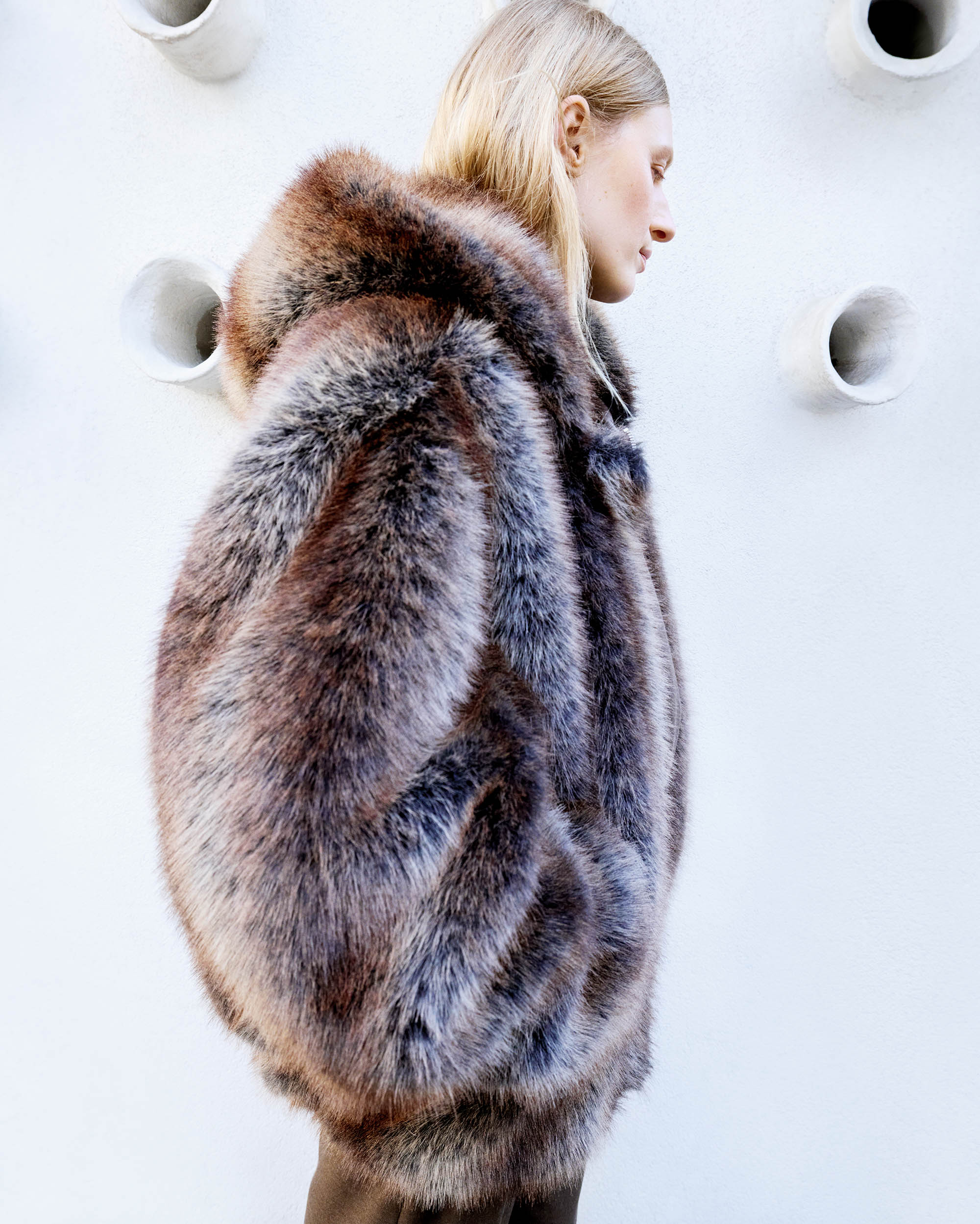 Adult female model poses in a brown and gray oversized fur coat against a plain white wall with sculptural elements in a fashion editorial.