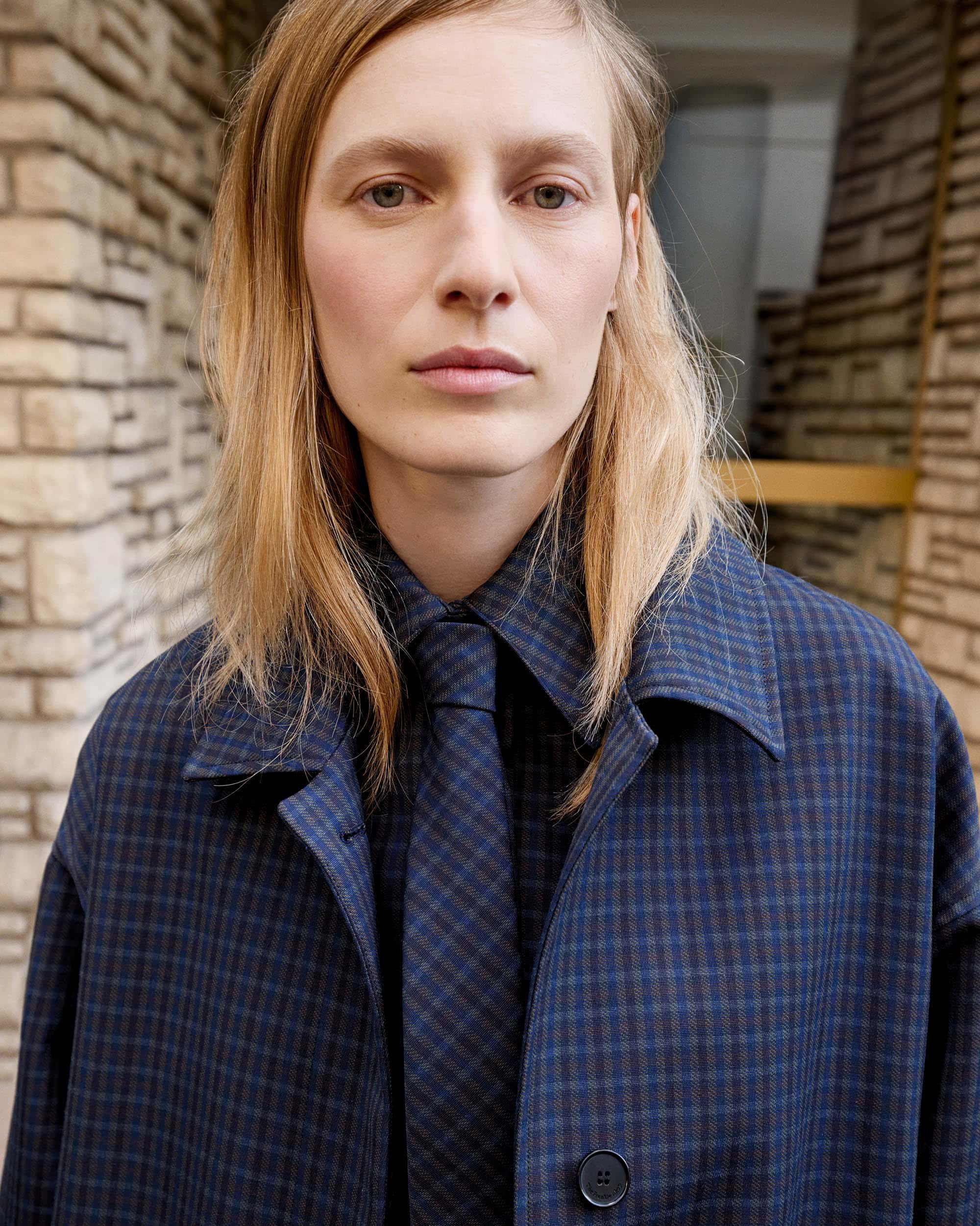 Close-up of a blonde woman in a blue plaid overcoat and tie, standing in front of a brick wall with a neutral, emotionless facial expression.
