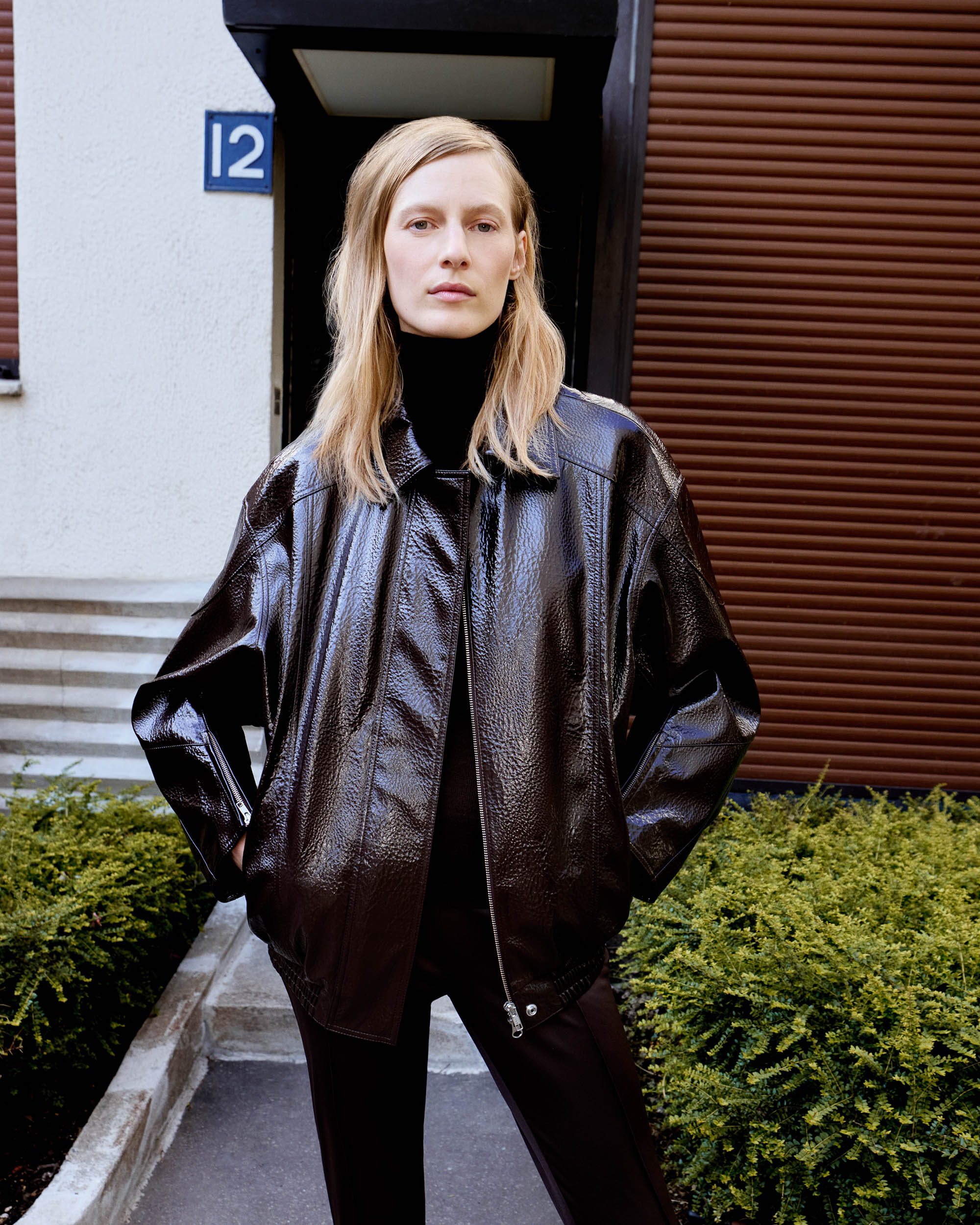 A blonde adult woman stands outdoors in a brown, high-shine jacket and black turtleneck, posing in front of a building and green shrubs.