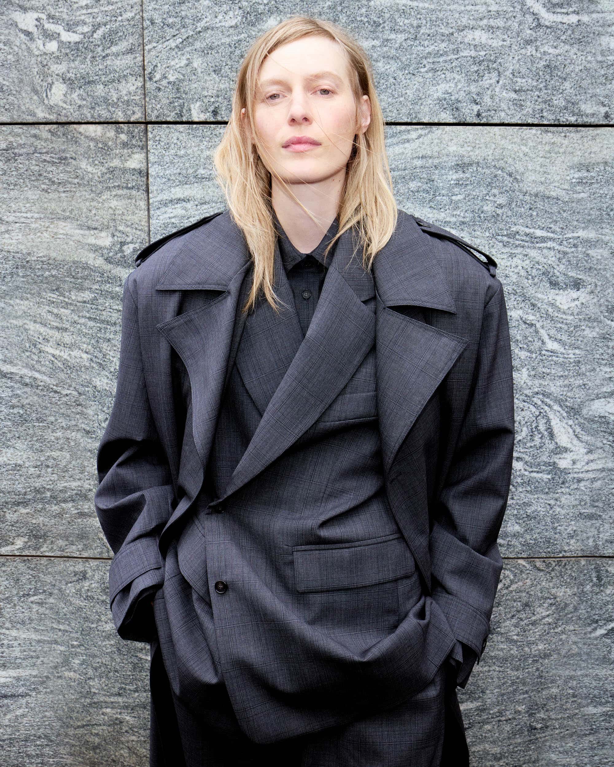 A woman with long blonde hair stands against a textured grey wall, wearing a dark grey suit with a double-breasted trench coat.
