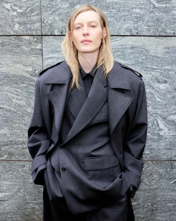 A woman with long blonde hair stands against a textured grey wall, wearing a dark grey suit with a double-breasted trench coat.