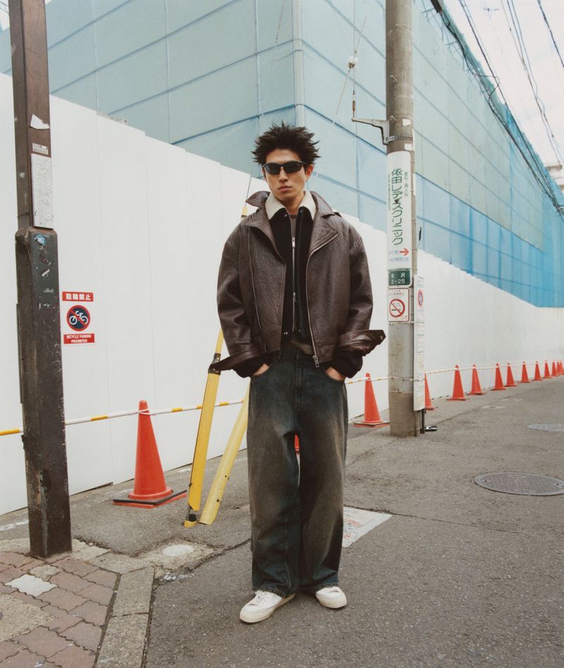 A young man poses in a brown leather jacket and baggy jeans on a city street with construction and traffic cones in the background.