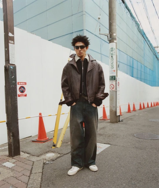 A young man poses in a brown leather jacket and baggy jeans on a city street with construction and traffic cones in the background.
