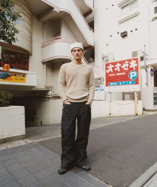 A young adult man stands on a street next to a building and signage, wearing a beige sweater and dark pants in an urban setting.