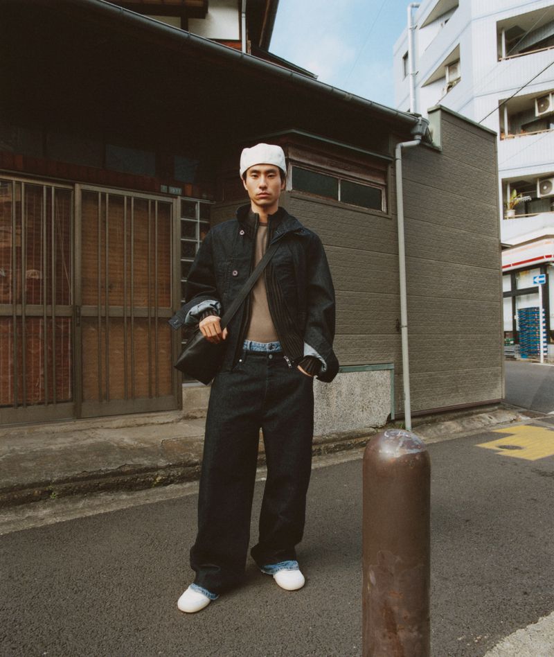 A Japanese man in a denim jacket and wide-leg pants, stands outside a traditional building on an urban street, holding a bag.