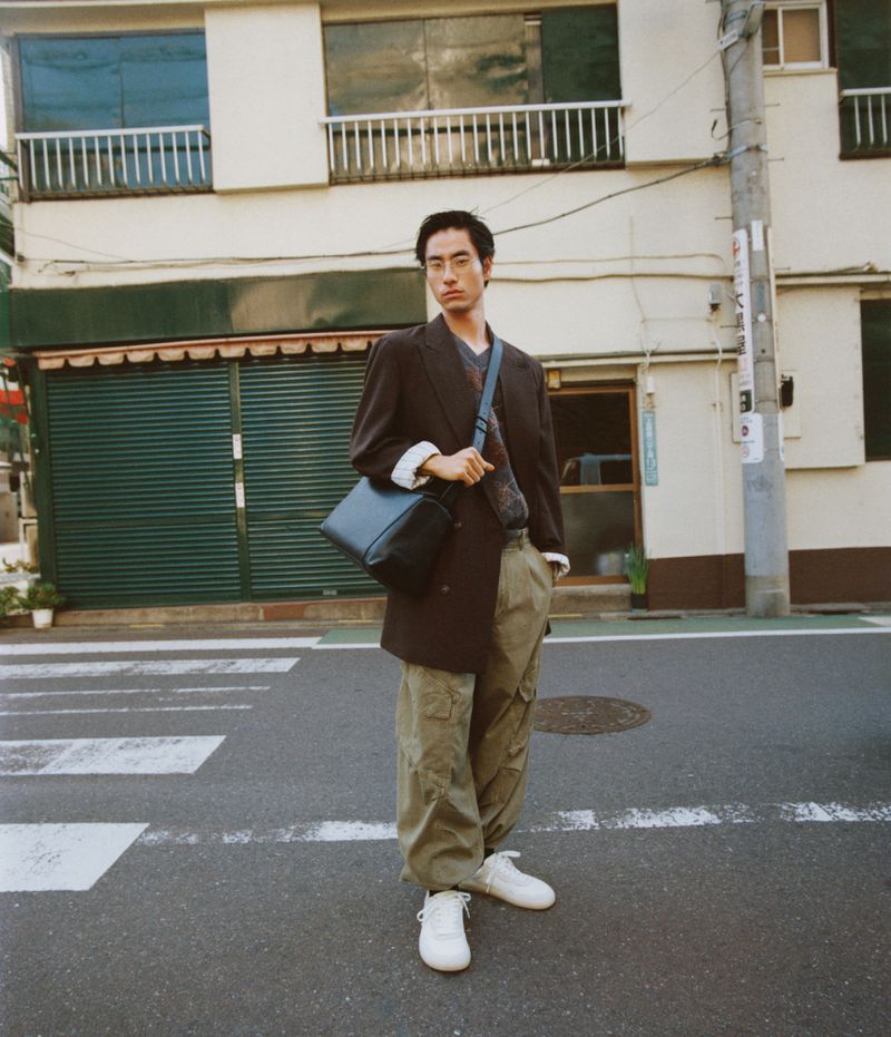 Stylish Asian male standing on a crosswalk in a brown blazer, tan cargo pants, and white sneakers, carrying a dark blue messenger bag.