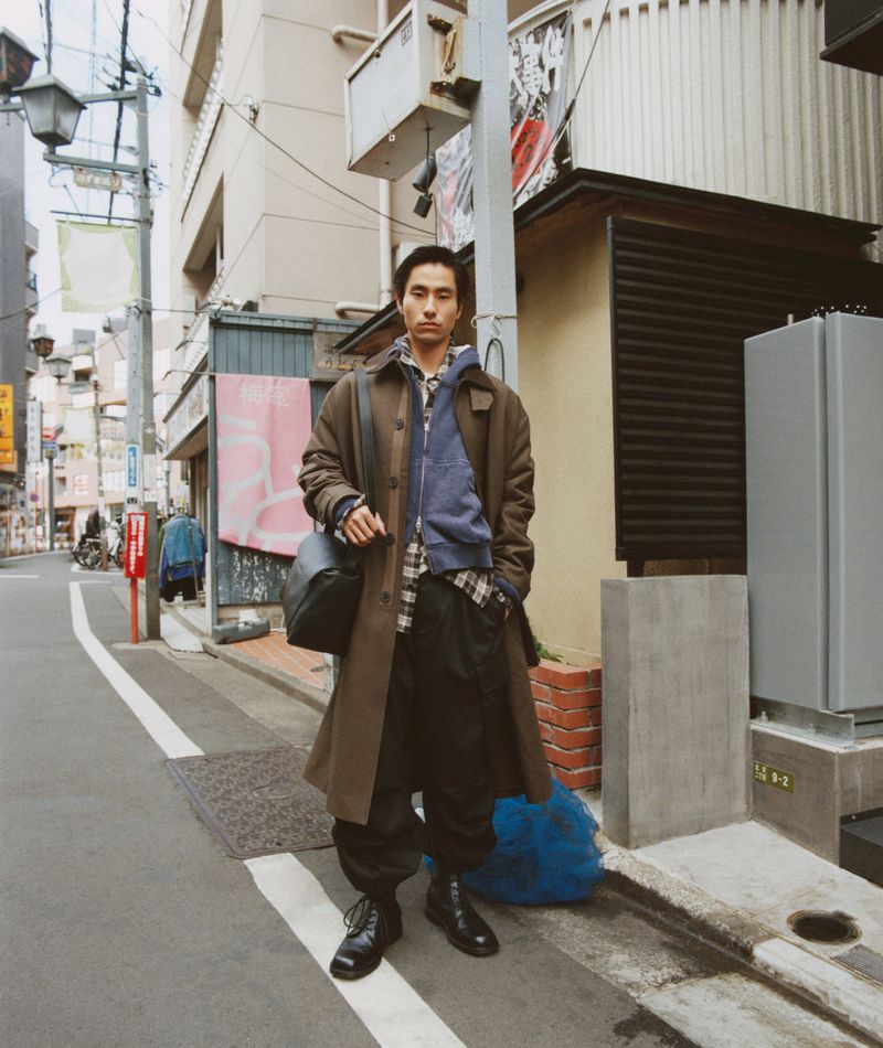 A male adult in outdoor urban street environment wearing a long brown coat, blue hoodie, plaid shirt, dark pants, and carrying a black bag.