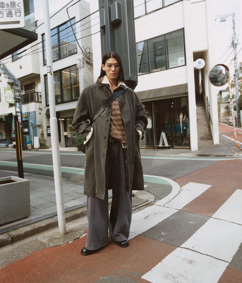 Adult male in casual streetwear poses on a crosswalk, wearing a dark coat, striped shirt, and wide-leg pants with buildings in the background.