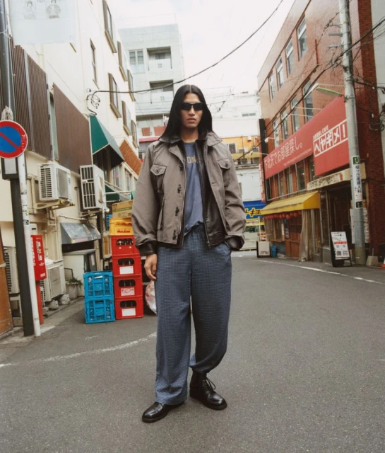 Man in jacket and plaid pants stands in a city street framed by buildings, overhead wires, stacked crates and muted daytime lighting.