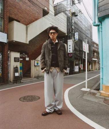 A man with sunglasses stands on a city street wearing a brown jacket, sweater vest, and baggy gray pants in an urban setting.