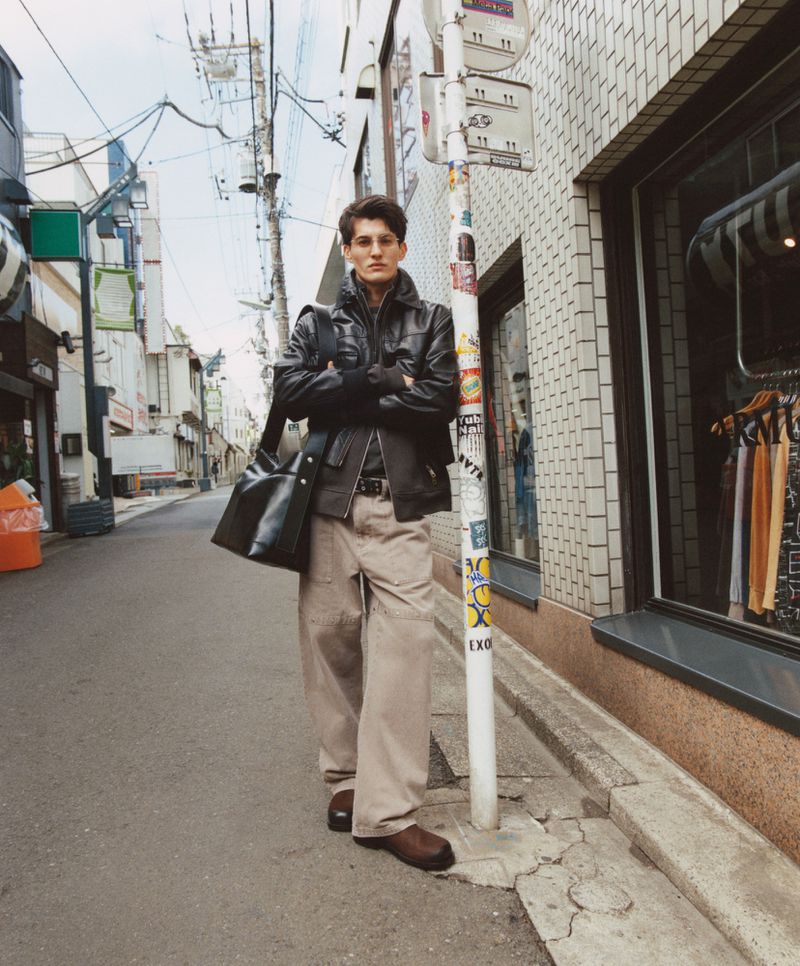 Male model in a black jacket and khaki pants stands on a city street near a bus stop, carrying a black bag with arms crossed.
