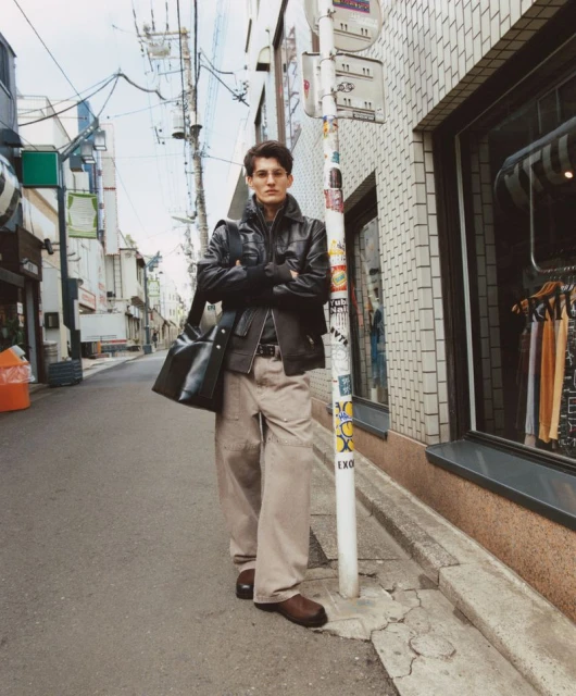 Male model in a black jacket and khaki pants stands on a city street near a bus stop, carrying a black bag with arms crossed.