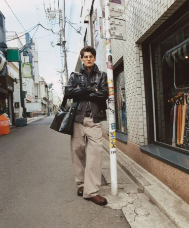 Male model in a black jacket and khaki pants stands on a city street near a bus stop, carrying a black bag with arms crossed.