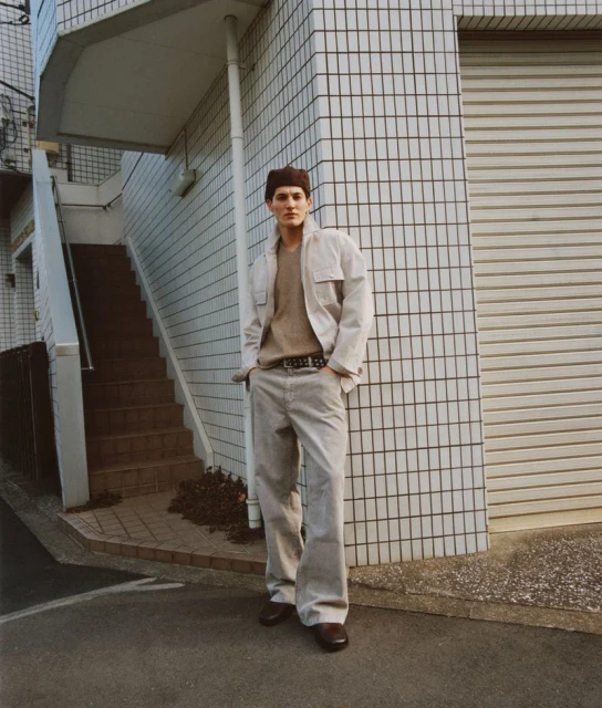 Man in a beige outfit stands against a white tiled building with a garage door and stairs, showcasing a relaxed pose outdoors.