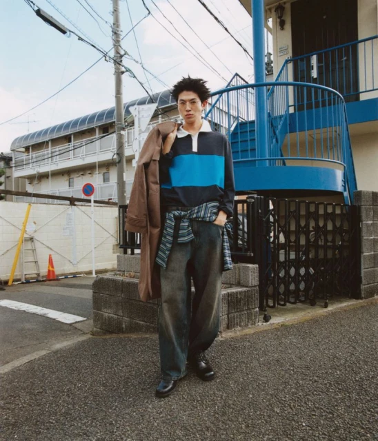 Man in striped shirt with coat slung over shoulder, standing on urban street near blue building. Fashion editorial outdoor setting.