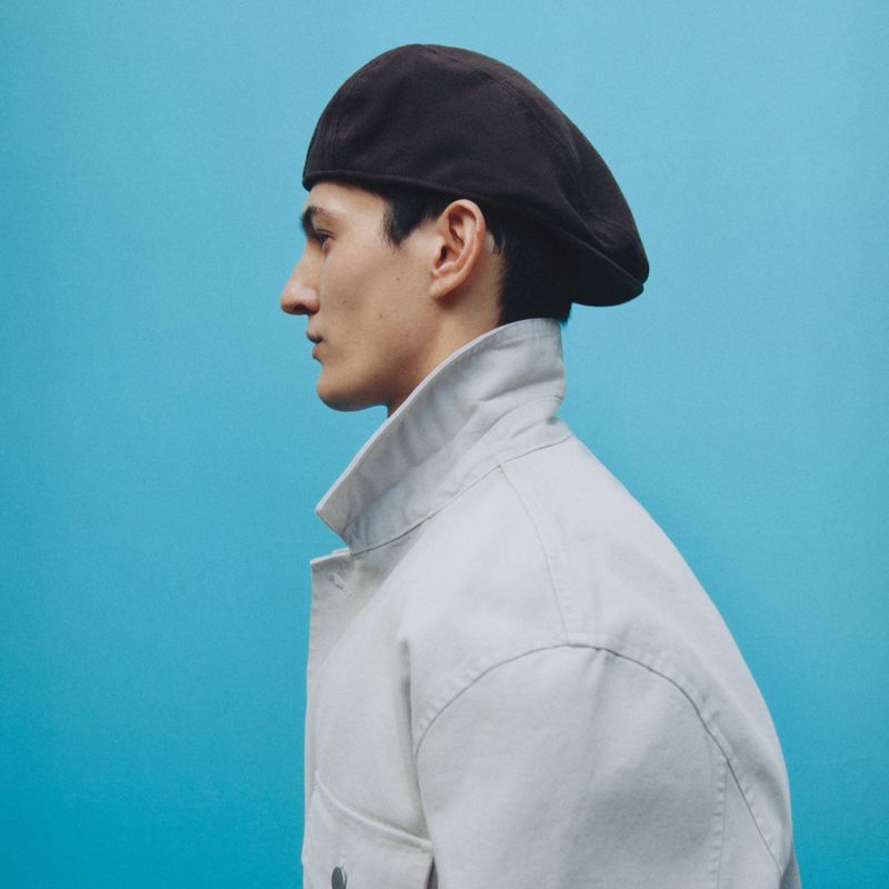 Profile of a young adult wearing a brown beret and a light grey jacket against a solid turquoise background in a studio portrait.