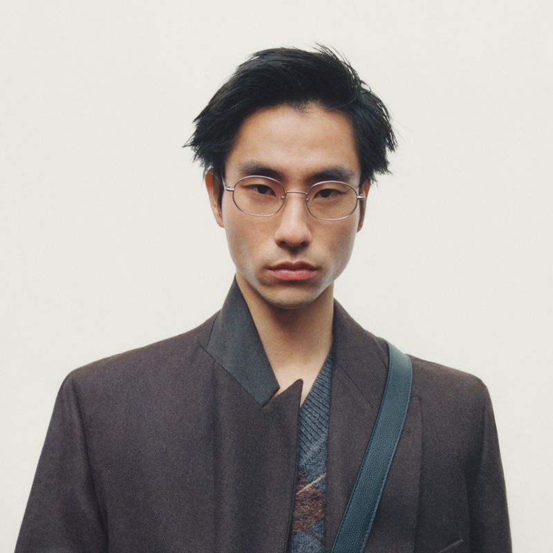 Portrait of a man wearing metal-frame glasses and a dark brown jacket against a plain light background