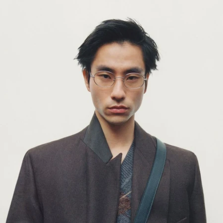 Portrait of a man wearing metal-frame glasses and a dark brown jacket against a plain light background