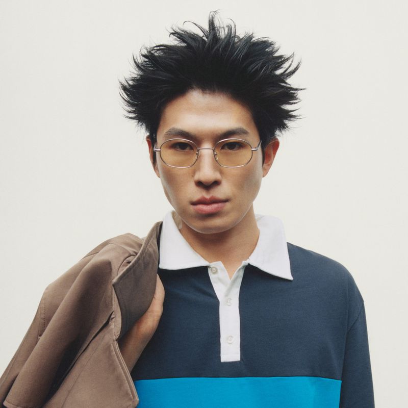 Portrait of a man wearing glasses and a color-blocked polo shirt holding a brown jacket over his shoulder against a plain background