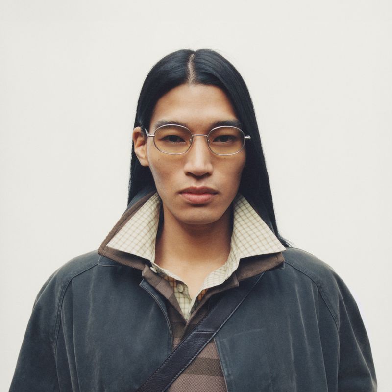 Headshot of an androgynous person with long, dark hair wearing glasses, a polo shirt, and a collared jacket against a white backdrop.