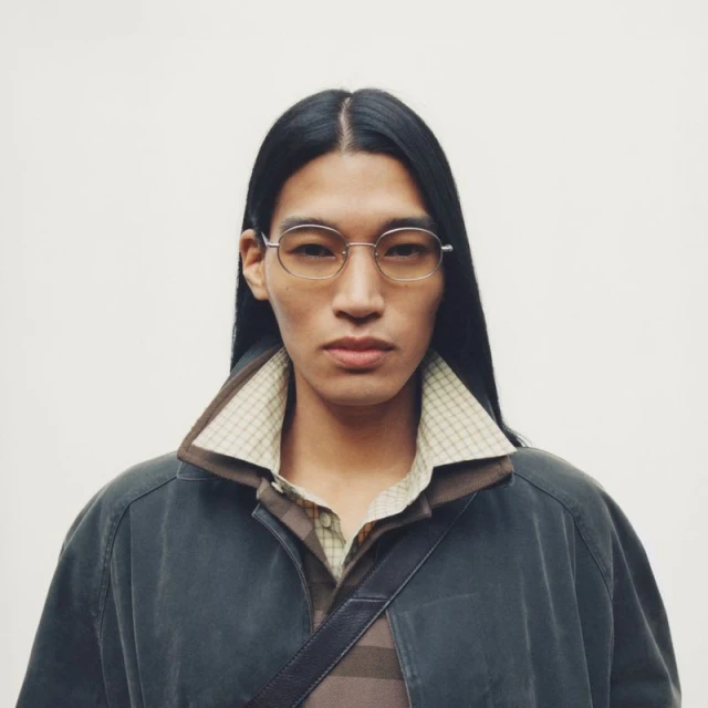 Headshot of an androgynous person with long, dark hair wearing glasses, a polo shirt, and a collared jacket against a white backdrop.