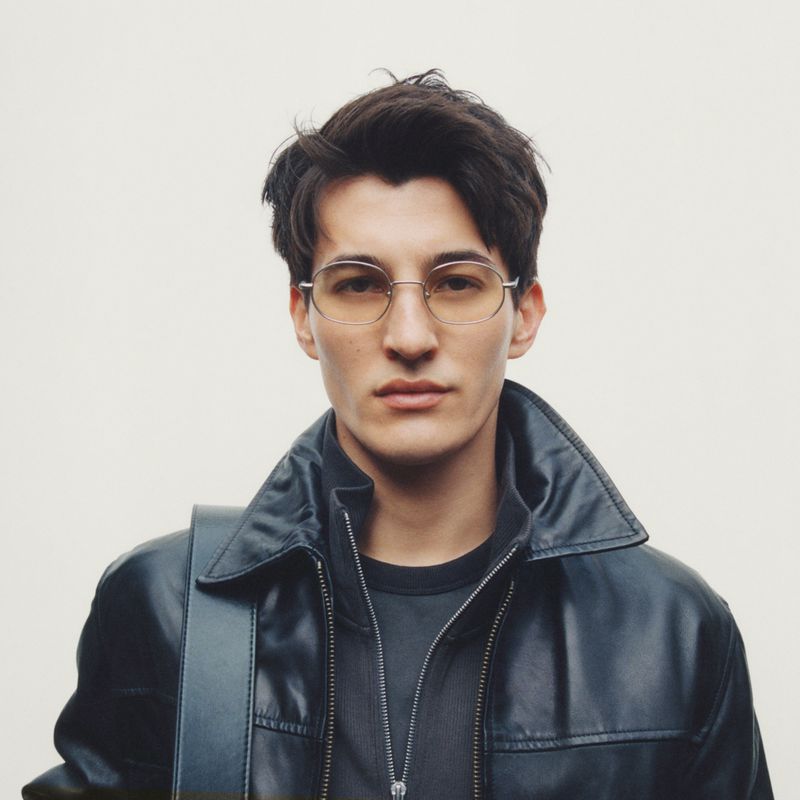 A young adult man wearing round eyeglasses and a leather jacket poses indoors against a light background, looking directly at the camera.