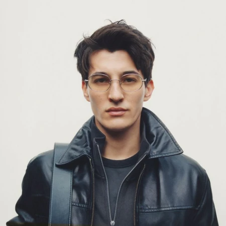 A young adult man wearing round eyeglasses and a leather jacket poses indoors against a light background, looking directly at the camera.