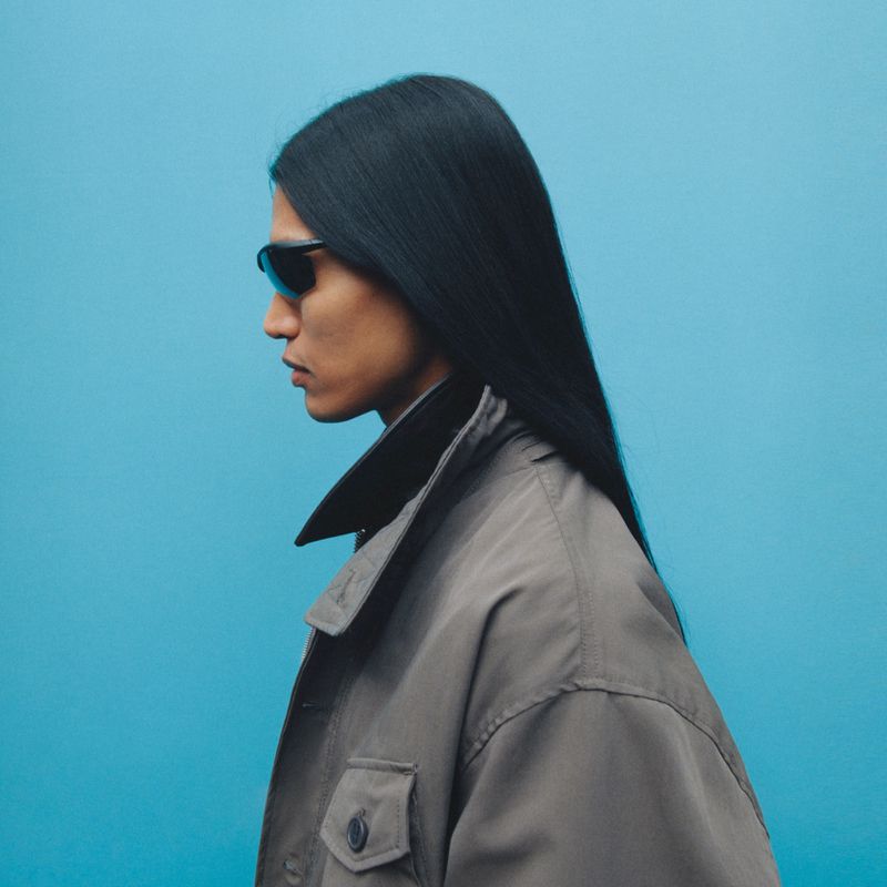 Profile of an adult with long black hair, wearing sunglasses and a gray jacket against a blue background in a studio shot.