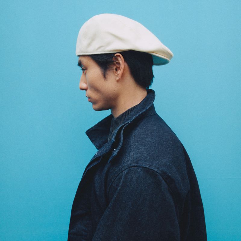 Profile view of a stylish Asian man wearing a cream cap and a dark blue denim jacket against a vibrant turquoise background.