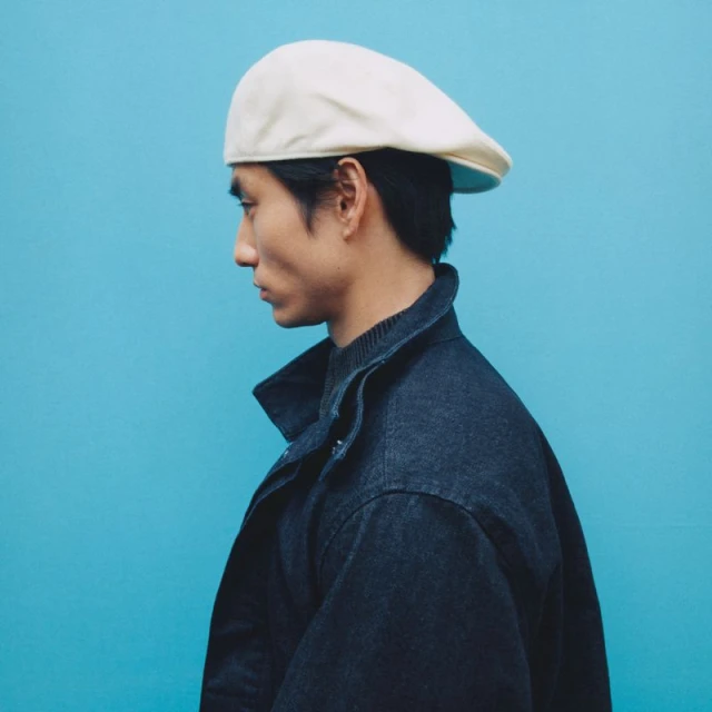Profile view of a stylish Asian man wearing a cream cap and a dark blue denim jacket against a vibrant turquoise background.