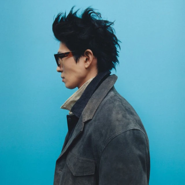 Side profile of an adult male with dark hair wearing glasses and a gray jacket against a light blue background in a studio setting.