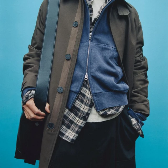 Fashionable menswear layers: olive-green coat, blue fleece jacket, and black pants. Stylish outfit with a shoulder bag against teal.