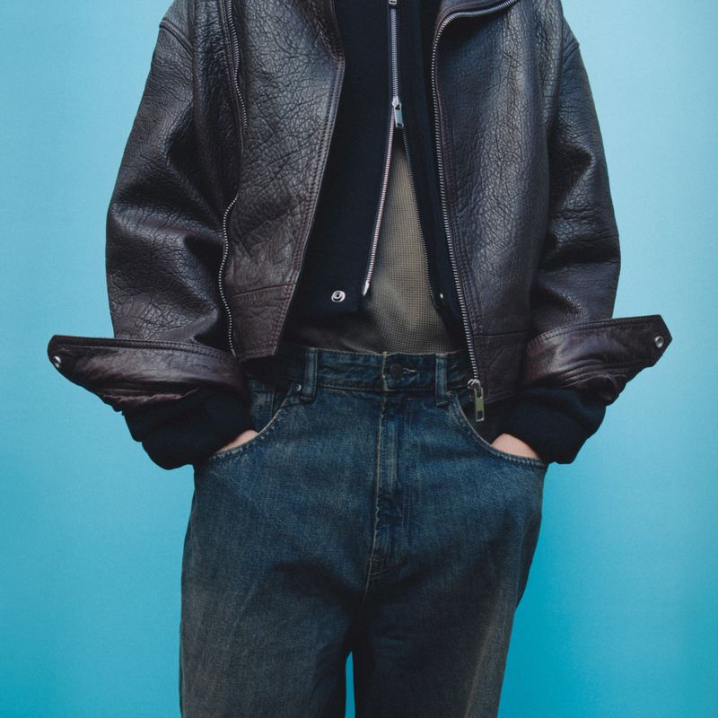 Person wearing a dark brown leather jacket with a zipper sweater underneath, posing in blue jeans against an aqua blue background.