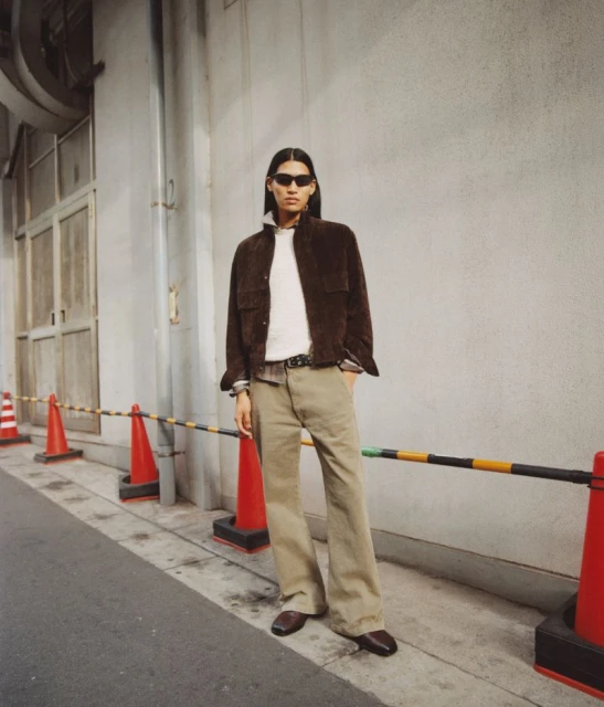 A person stands in front of a wall wearing sunglasses, a brown jacket, and khaki pants, complemented by traffic cones and a barricade.